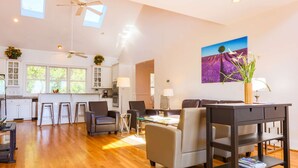 A spacious living area with a wooden floor, a dining table, and a sofa. There are two ceiling fans, a kitchen with white cabinets, and a large window.