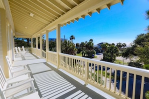 A covered porch with white railings and chairs, overlooking a street with palm trees and buildings. - Steps to Beach. Relax in Dog Friendly Ocean View Home, 2 Master Suites & Porch (Folly Beach)