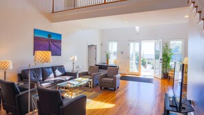 A spacious living room with a high ceiling, a black leather sofa, a glass coffee table, and a view of the outdoors through large windows.