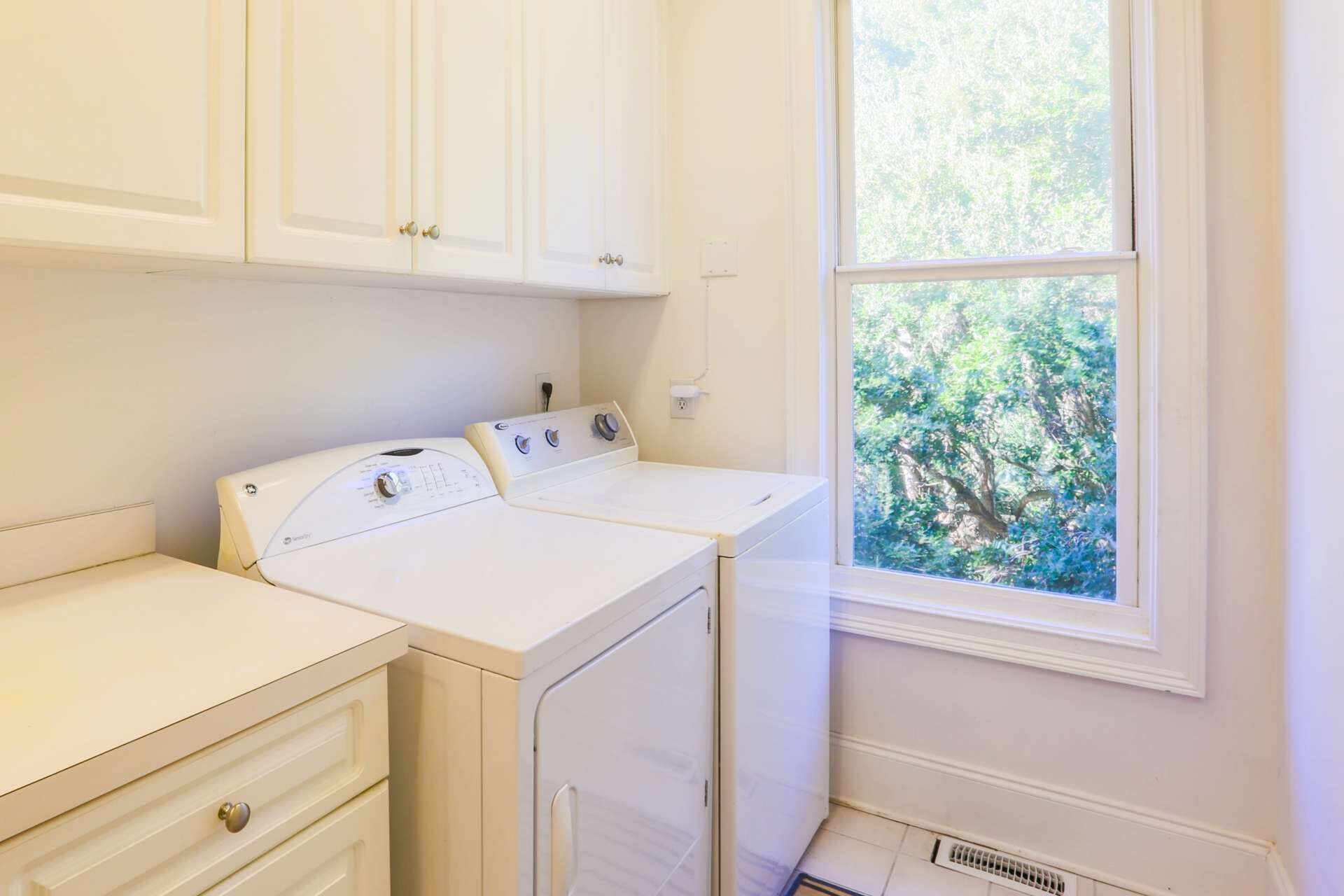 A laundry room with two white washers and dryers, a small cabinet, and a window with a view of trees.