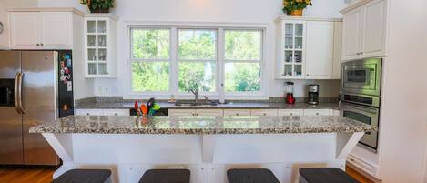 Una cocina moderna con armarios blancos, encimeras de granito, una isla central con taburetes, un refrigerador, un horno y un microondas.