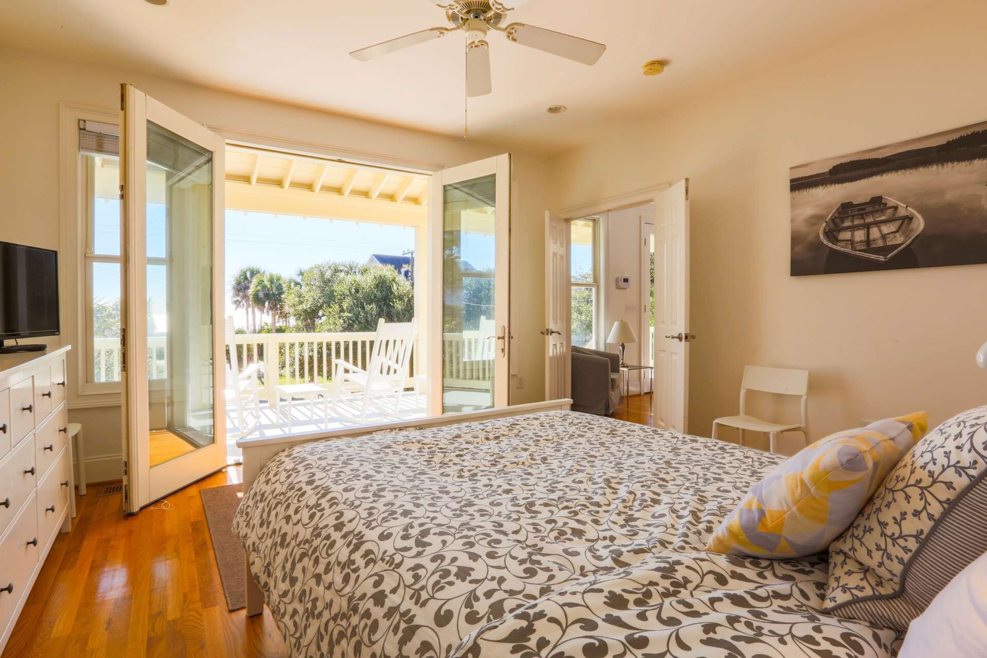 A bedroom with a bed, a television, a ceiling fan, and a balcony with a view of trees and a building.