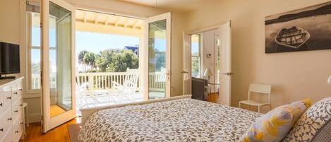 A bedroom with a bed, a television, a ceiling fan, and a balcony with a view of trees and a building.