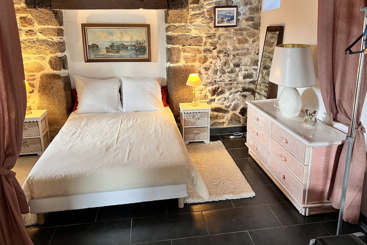 Une chambre avec un mur en pierre, un lit avec une literie blanche, une table de chevet avec une lampe, une commode et un tableau encadré au mur.