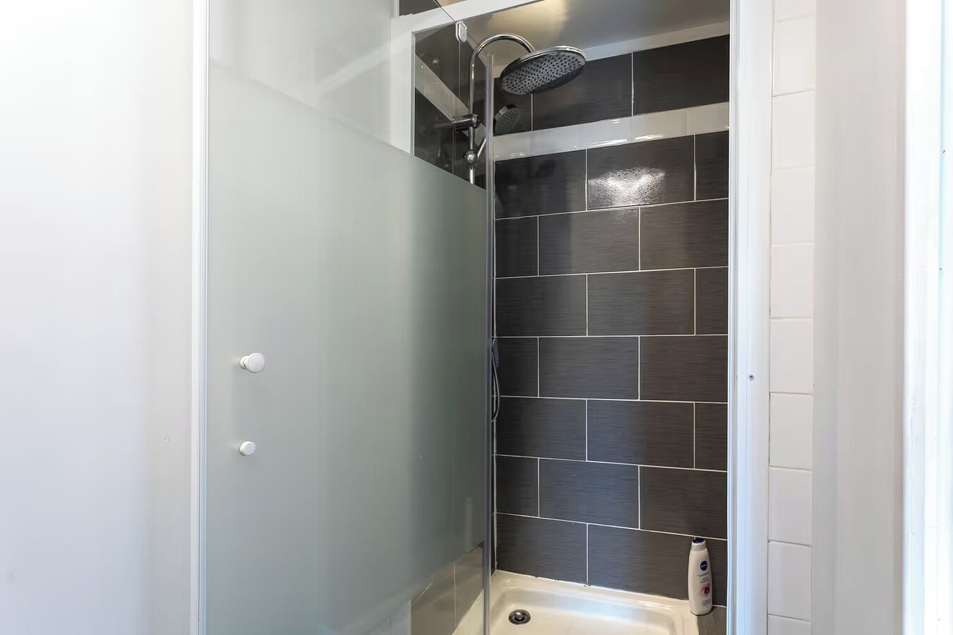 Een douche met een handdouche, een wit bad en donker betegelde wanden.
