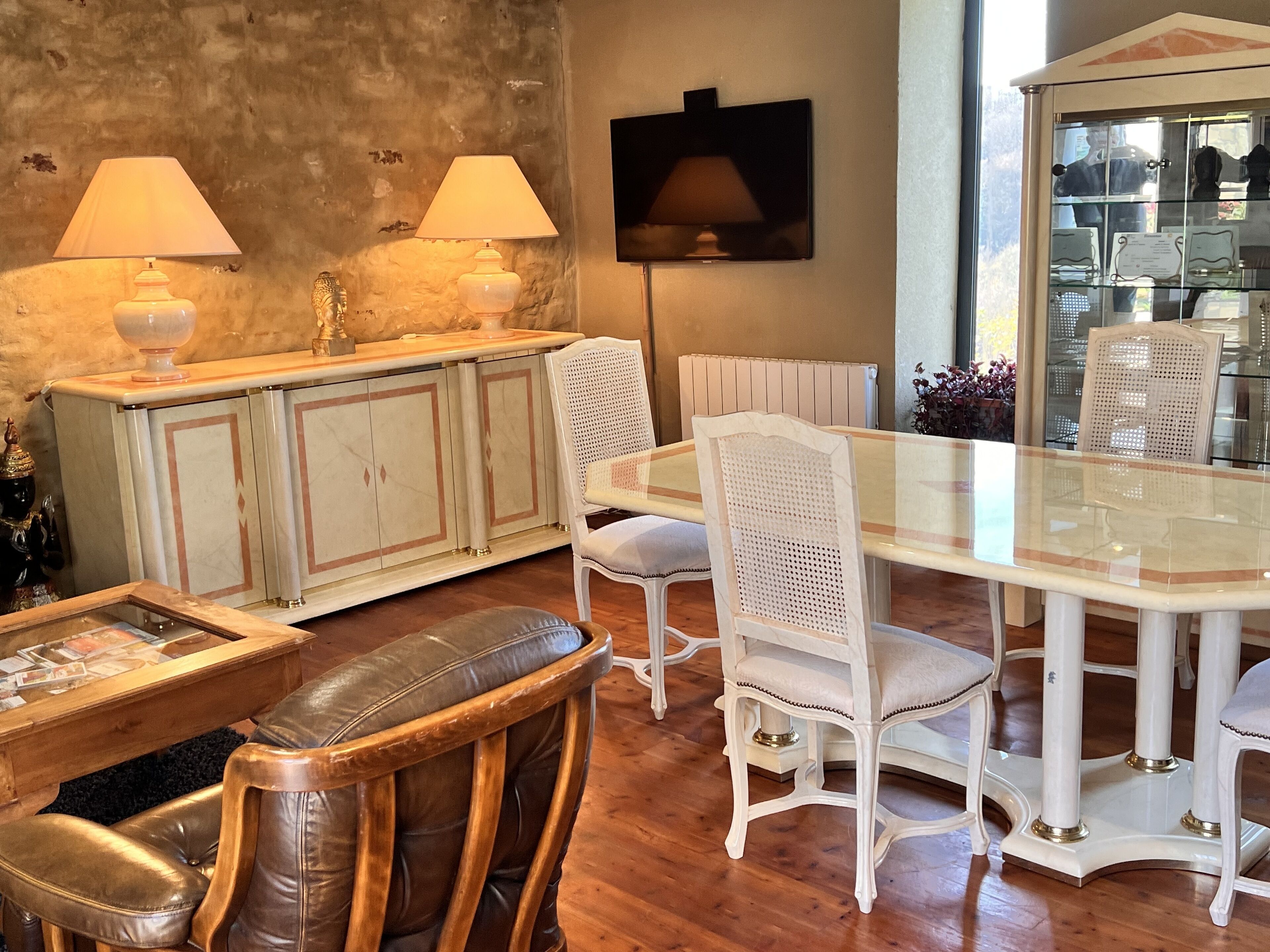 Un coin repas avec une table à plateau en verre, des chaises à revêtement blanc et un canapé en cuir. On y trouve également un meuble blanc orné d’objets décoratifs et un téléviseur fixé au mur.