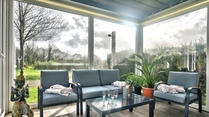 Un patio iluminado por el sol con tres sofás grises, una mesa de vidrio y plantas en macetas.