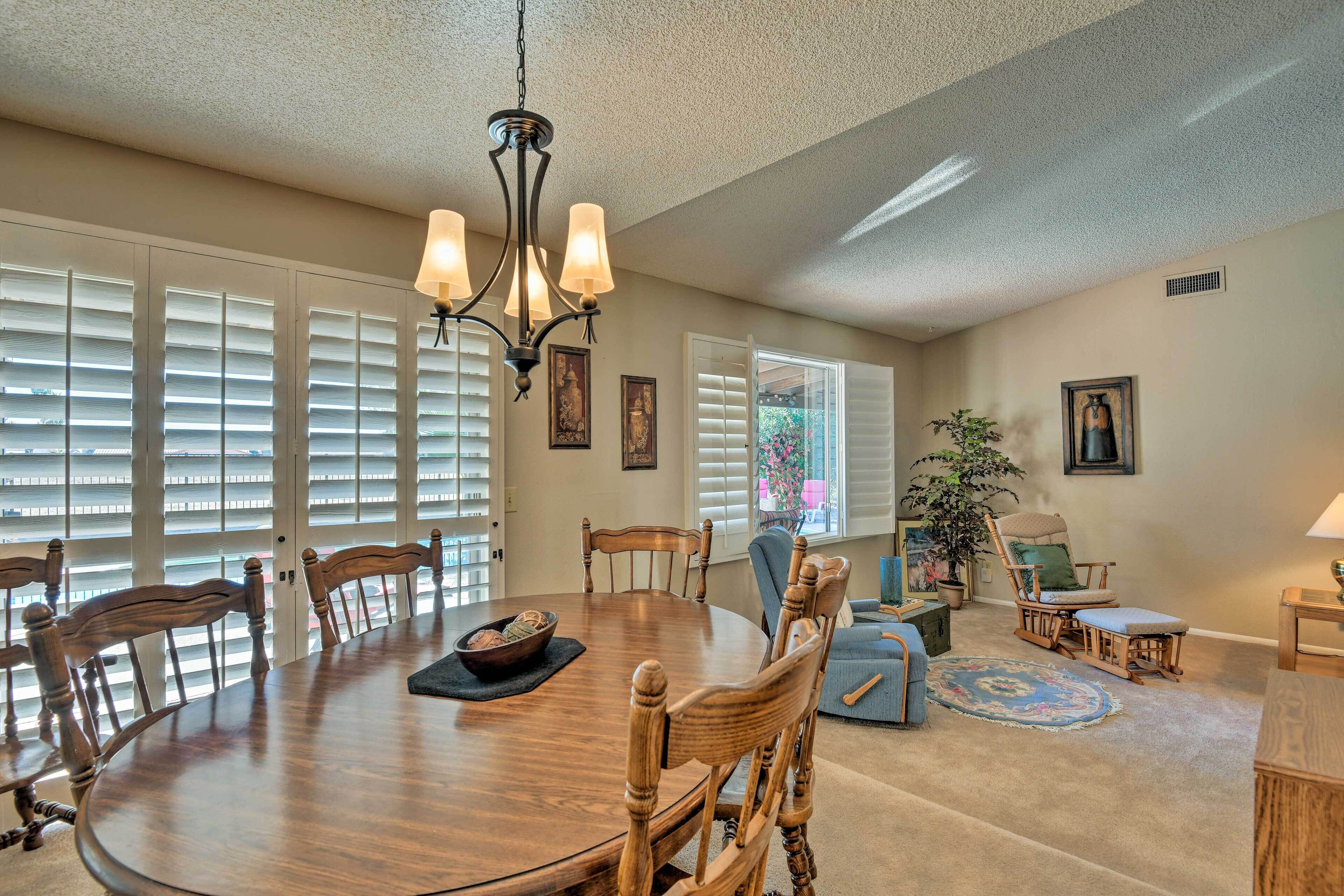 A dining area with a round table, wooden chairs, a chandelier, and large windows with blinds.