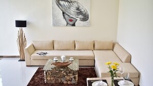 Een moderne woonkamer met een beige bank, een glazen salontafel met een houten kom, een eethoek met een vaas vol gele bloemen, en een schilderij aan de muur.