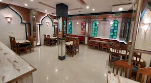 A dining area with wooden tables and chairs, a central pillar, and arched windows with colored glass. - Hotel Reeve Inn (Jaipur)
