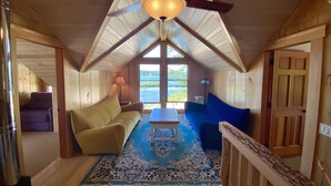 A cozy attic room with a sloped ceiling, wooden walls, a ceiling fan, a large window with a view of a body of water, a blue and beige patterned rug, and a wooden bench. - Booking for next summer. (Center Ossipee)