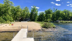 A wooden dock leading to a sandy beach with clear water, surrounded by lush green trees and a house in the background. - Booking for next summer. (Center Ossipee)