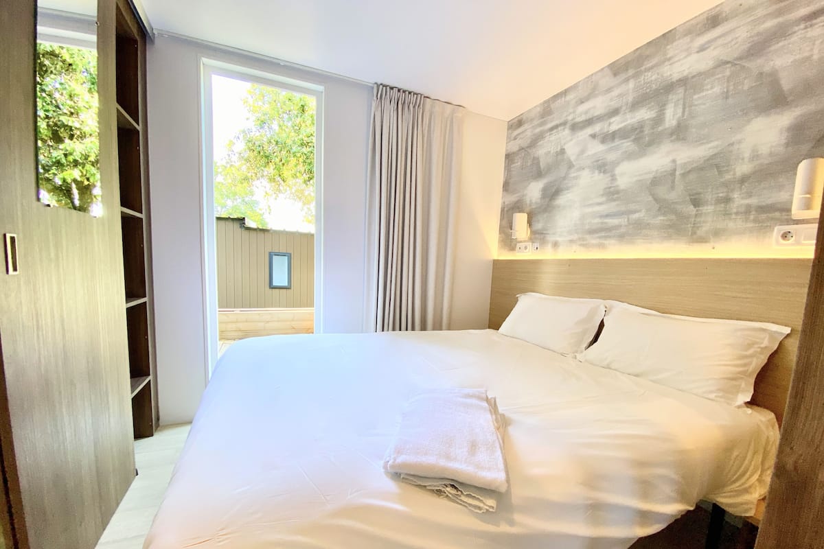 A modern hotel room with a large bed, a wardrobe, and a view of the outdoors.