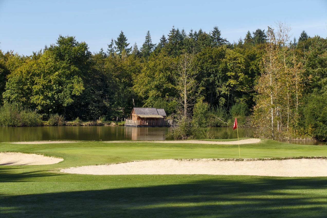 A golf course with a thatched-roof building near a lake, surrounded by dense trees.