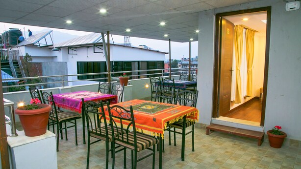 Un área de patio al aire libre con sillas y mesas de metal, plantas en macetas y vistas a edificios en el fondo.