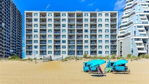A multi-story hotel with balconies, a sandy beach in the foreground, and beach chairs and umbrellas. - 🏖️ Delightful Ocean Front Unit - Breathtaking Views - Perfect for Families! 🏖️ (Ocean City)