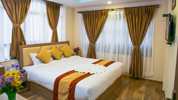 Una habitación de hotel con una cama grande, cortinas, una mesita de noche, un jarrón con flores y una lámpara en el techo.