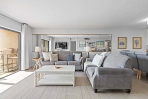 A modern living room with a sofa, coffee table, and chairs. There is a TV mounted on the wall and artwork displayed.