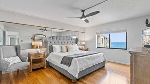 A bedroom with a large bed, a ceiling fan, a nightstand, a lamp, a chair, and a window with a view of the ocean.