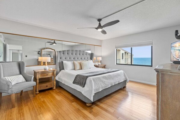 Une chambre à coucher avec un grand lit, un ventilateur de plafond, une table de chevet, une lampe, une chaise et une fenêtre donnant sur l’océan.