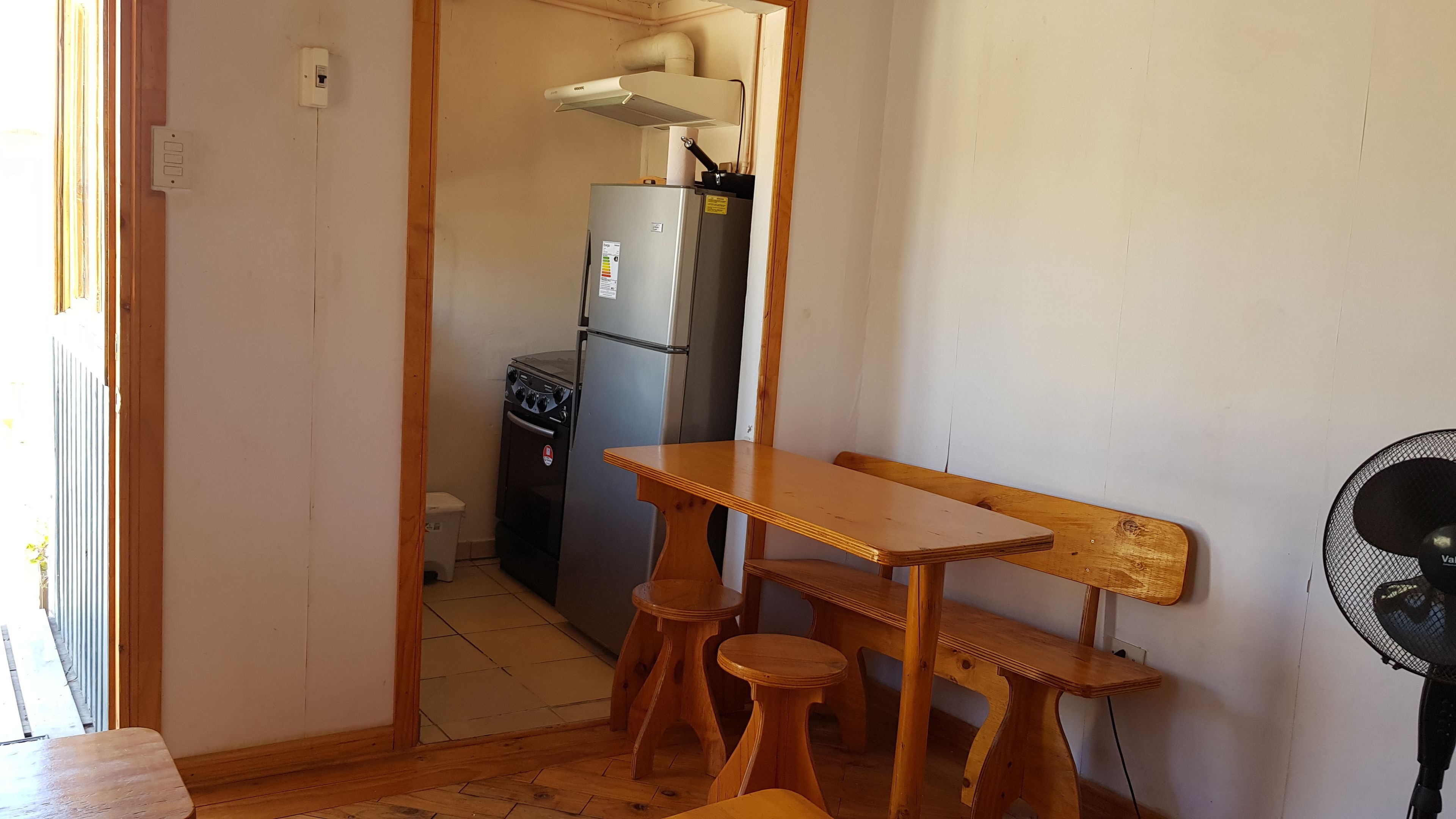A small kitchenette with a refrigerator, stove, and sink. There is a wooden table with two stools and a fan.