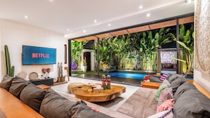 A modern living room with a large TV displaying 'NETFLIX', a wooden coffee table, a sofa, and a view of a pool with tropical plants.