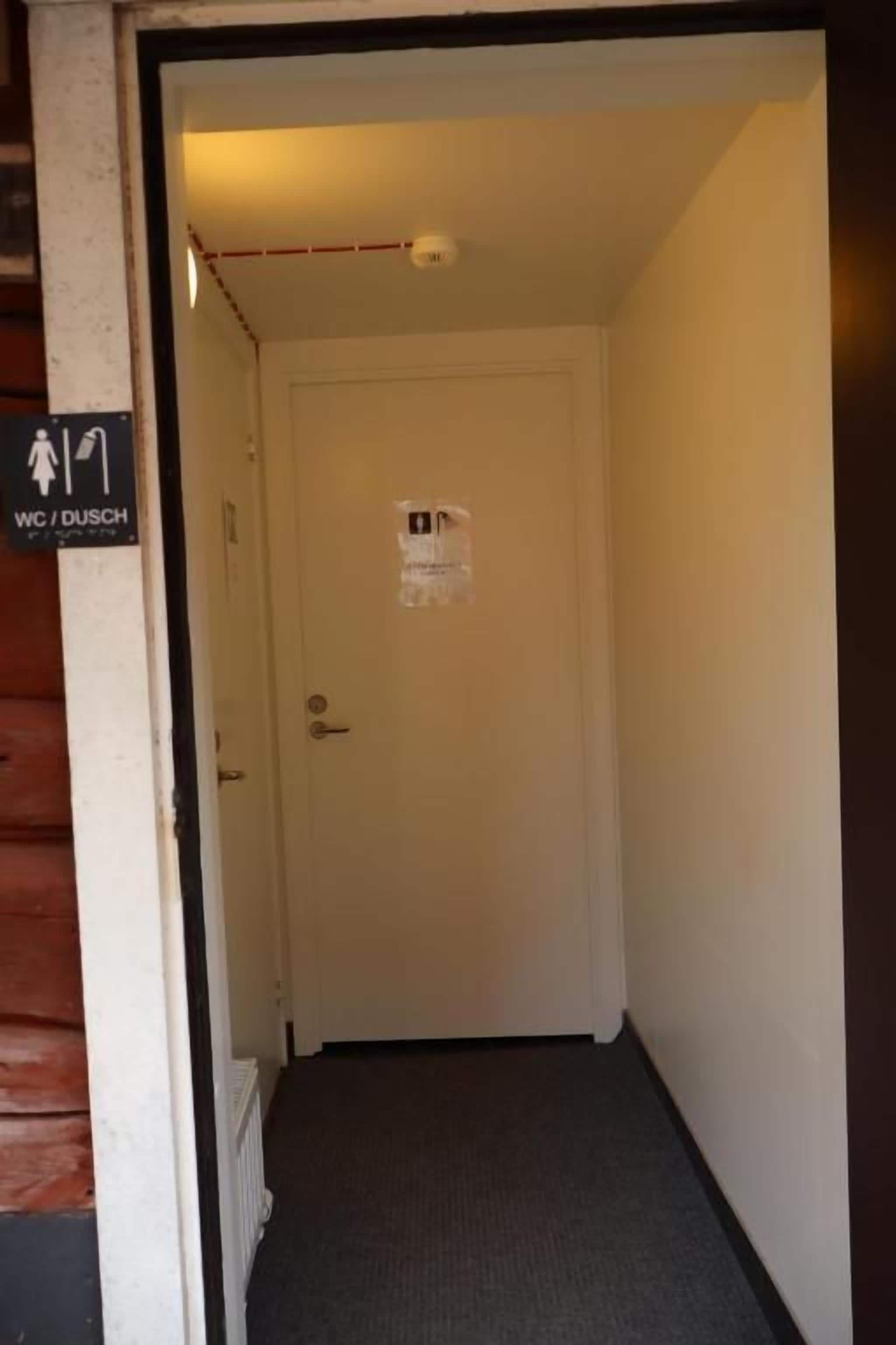 A narrow hallway with a door at the end, a sign indicating a women's restroom, and a fire alarm on the ceiling.