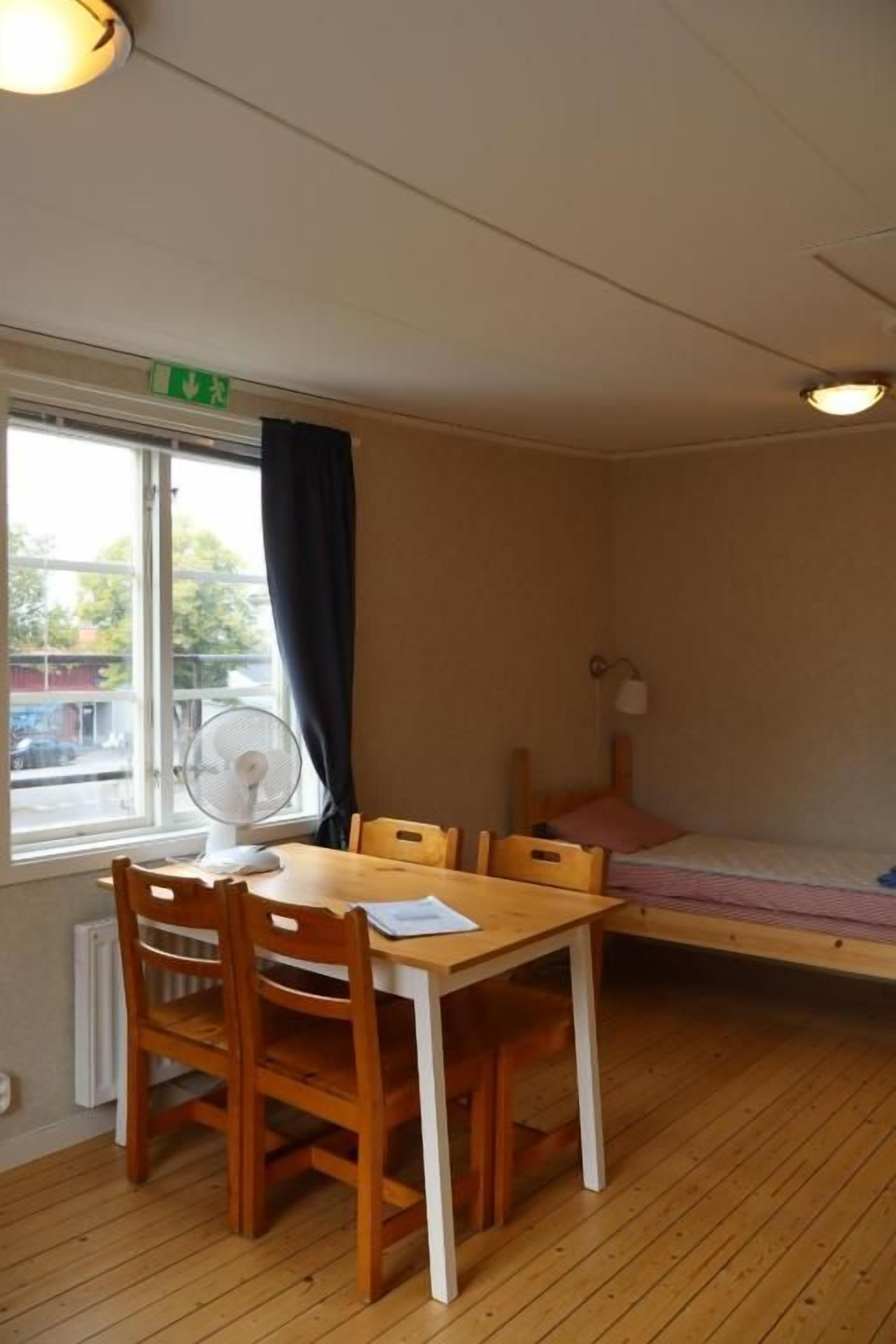 Una habitación con una mesa de madera y sillas, una cama, una ventana con cortinas y un ventilador de techo.