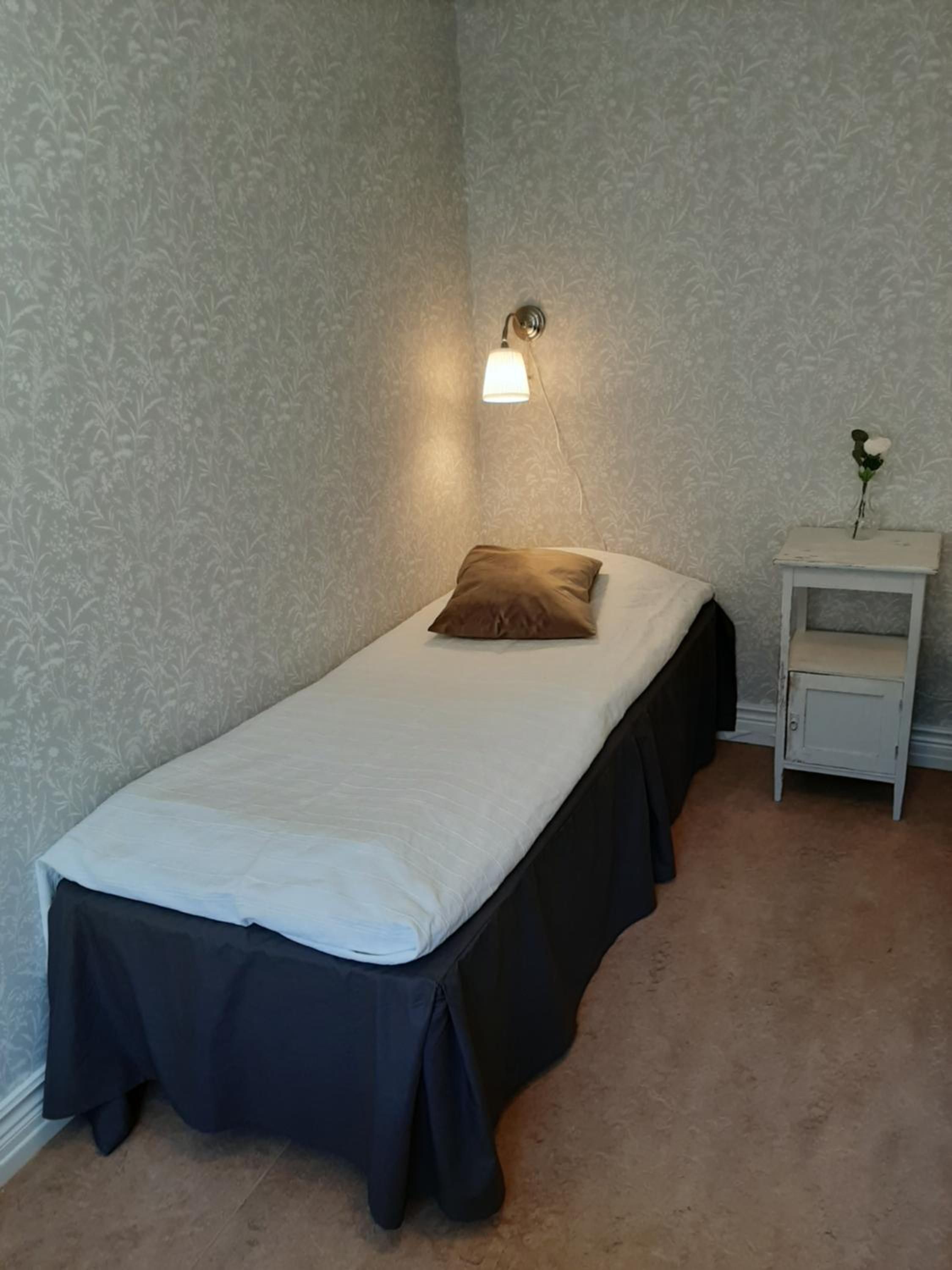 A single bed with a white mattress and dark skirted legs, a bedside lamp, and a small nightstand with a vase of flowers.