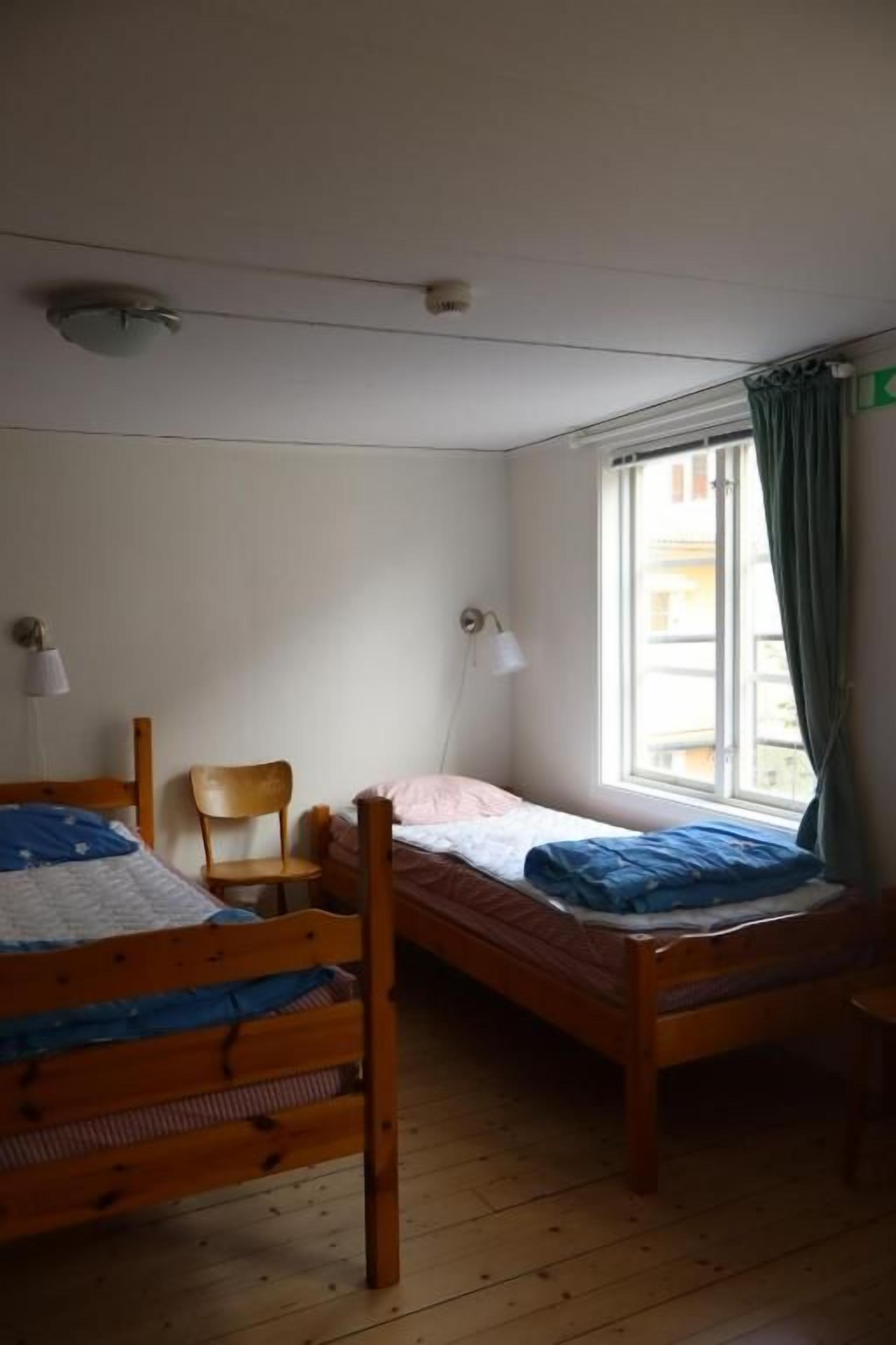 Quarto com duas camas de madeira, uma cadeira, uma janela com cortinas e uma luminária fixada na parede.