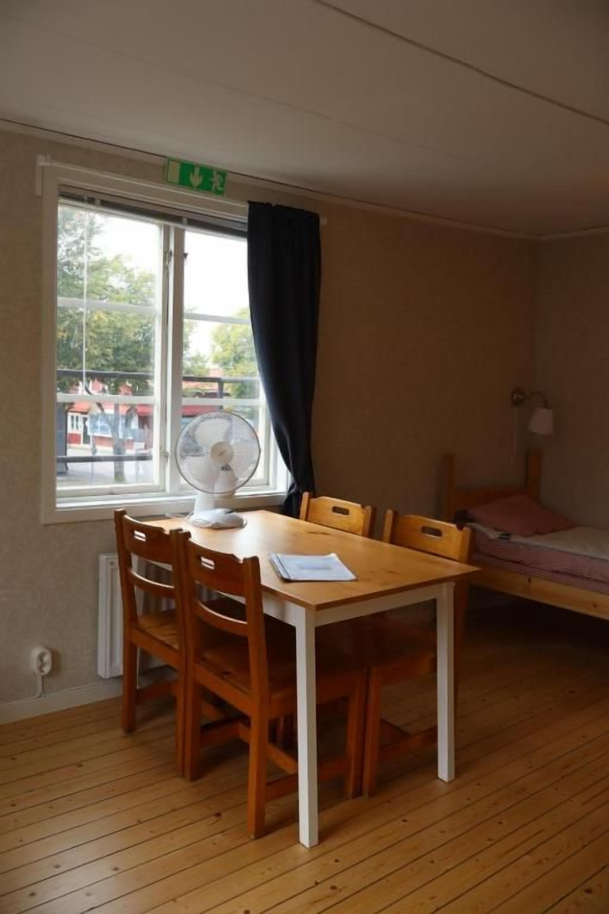 A room with a wooden floor, a table with chairs, a bed, a window with a curtain, and a fan.