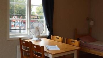 A room with a wooden floor, a table with chairs, a bed, a window with a curtain, and a fan.