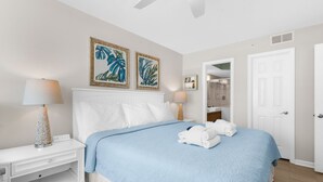Quarto de hotel com cama, duas luminárias na parede, mesa de cabeceira, porta branca e banheiro visível através de uma porta aberta.