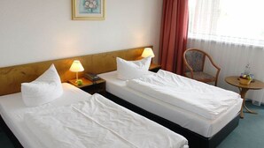 Een hotelkamer met twee eenpersoonsbedden, een houten hoofdbord, een nachtkastje met een lamp, een klein rond tafeltje met een mand, en een stoel.