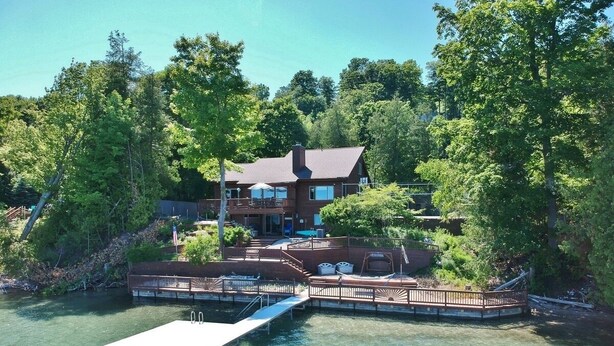 A lakeside property with a dock, a covered patio, and a large house surrounded by trees. - Luxury Bayfront Retreat on Grand Traverse Bay with Private Beach and Sport Court (Suttons Bay)