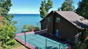 A tennis court adjacent to a wooden building with a view of the water. - Luxury Bayfront Retreat on Grand Traverse Bay with Private Beach and Sport Court (Suttons Bay)