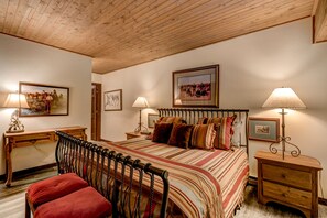 Um quarto aconchegante com uma cama de madeira, duas mesas de cabeceira com abajures, uma escrivaninha e uma cadeira.