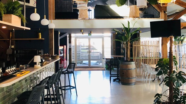 A spacious bar area with high ceilings and exposed beams, featuring hanging woven light fixtures and a variety of seating options. - See Breizh Camping La Ville Huchet (Saint-Malo)