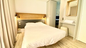 A hotel room with a large bed, wooden headboard, bedside lamps, a mirror, and a small seating area. - See Breizh Camping La Ville Huchet (Saint-Malo)