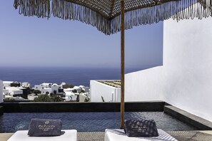 A poolside area with a view of the sea and white buildings, a wooden umbrella, and two towels with a logo.