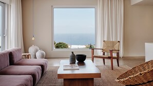 A modern living room with a large window, a sofa, a chair, and a coffee table. - Andronis Arcadia Hotel (Santorini)