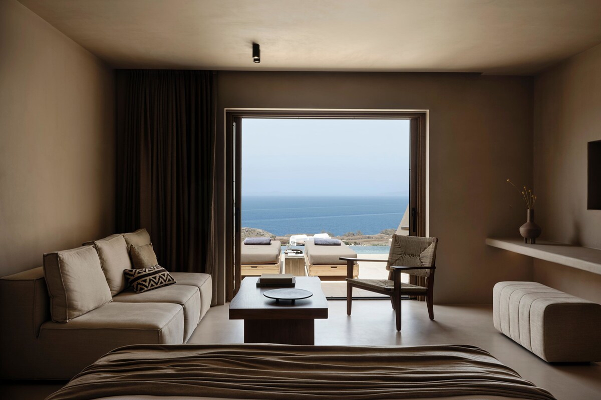 Habitación de hotel moderna con una cama grande, un sofá, una silla y una mesita pequeña. Se ve el océano a través de un ventanal amplio.