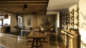 A spacious room with a wooden dining table, chairs, a hammock, and a wall-mounted shelf with plants. - Andronis Arcadia Hotel (Santorini)