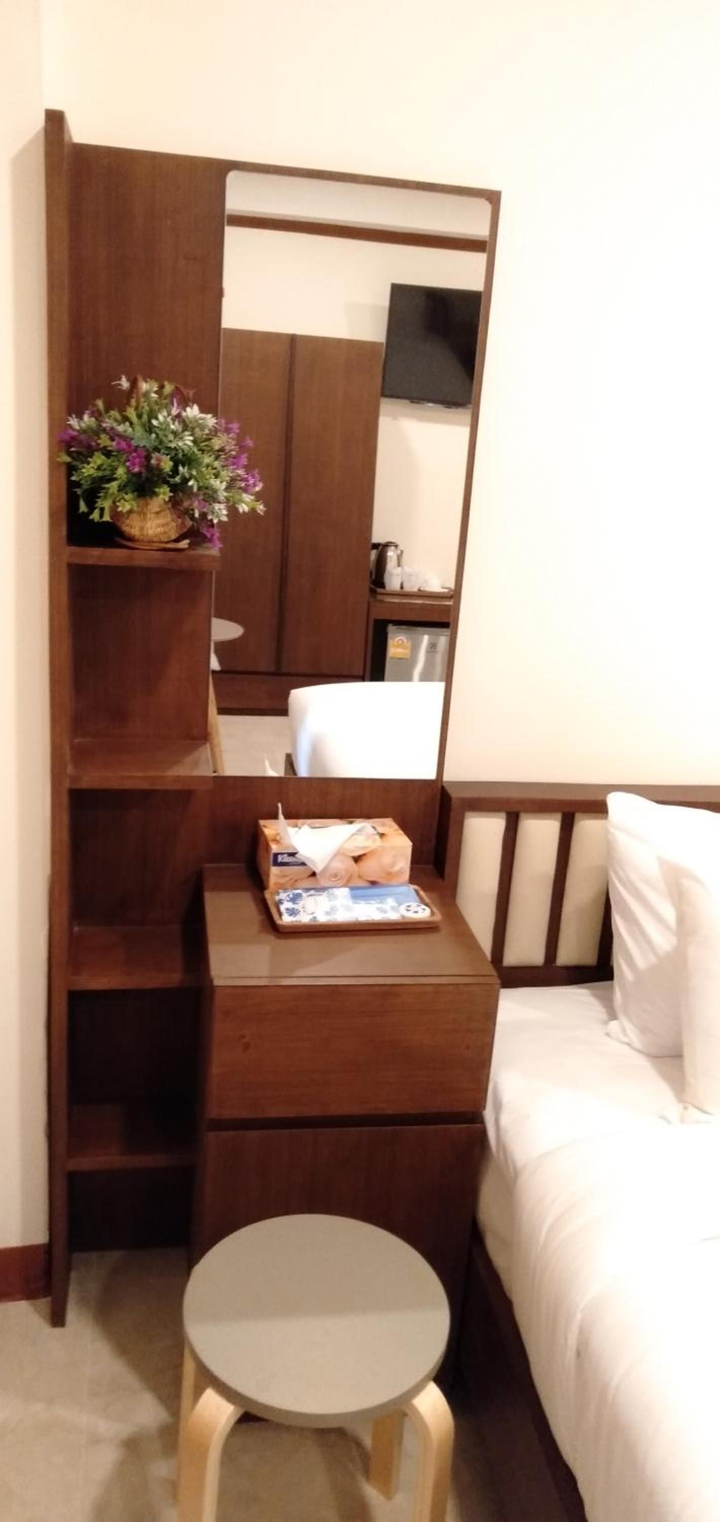 A hotel room with a bed, a bedside table with a mirror, a small stool, and a shelf with a plant.