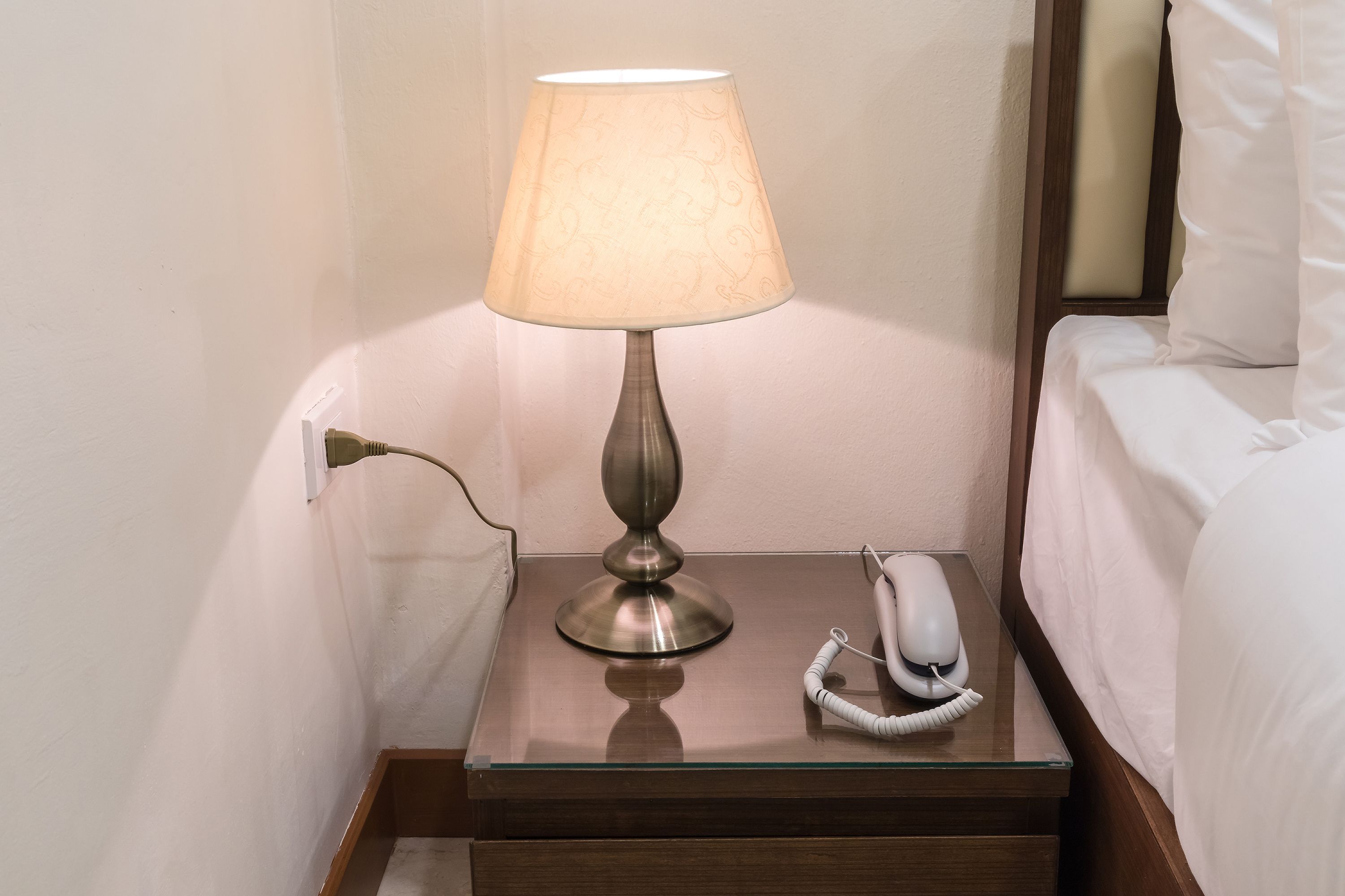 A bedside table with a lamp, a telephone, and a bedside drawer.