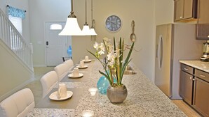 A dining area with a long table, white chairs, a vase with flowers, and a clock on the wall.
