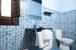 Un bany amb rajoles de mosaic blaus, un vàter blanc, un lavabo amb mirall i un suport per a tovalloles.