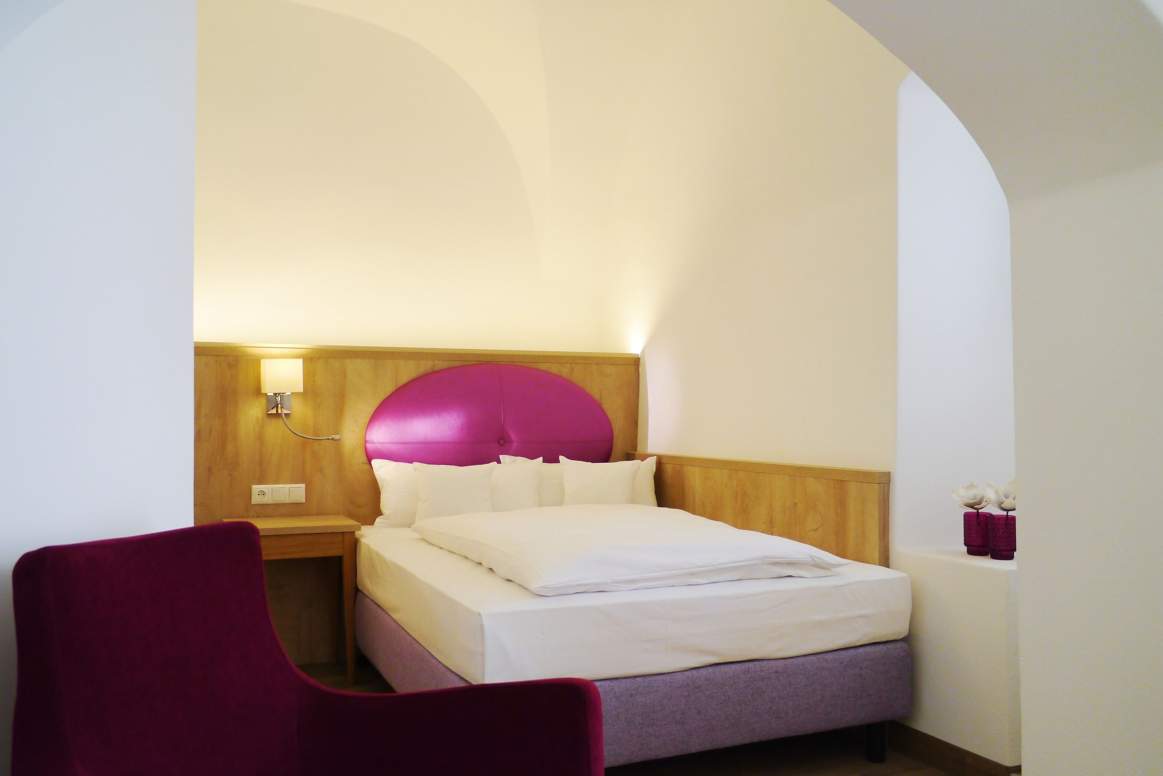 Ein Hotelzimmer mit einem Bett, einem Holzkopfteil, einem Nachttisch, einem roten Sessel und einer Wandlampe.