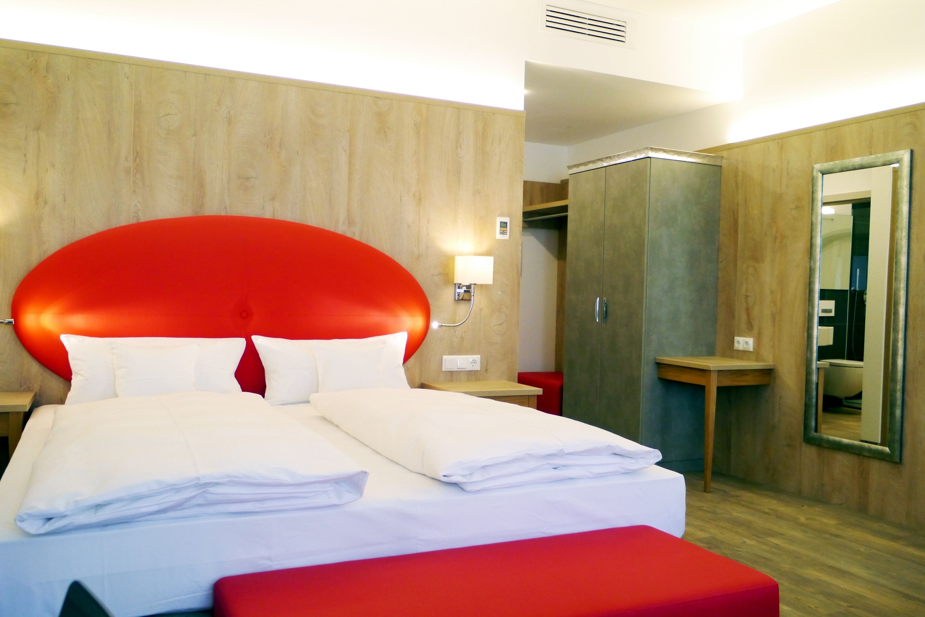 Ein Hotelzimmer mit einem großen Bett, einem roten Kopfteil, einer Holzwand, einem Kleiderschrank und einem Spiegel.