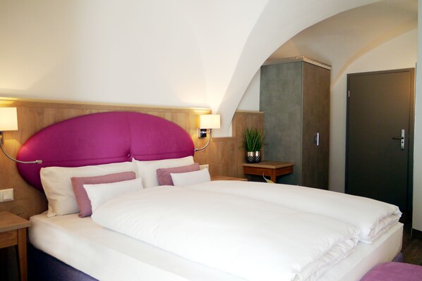 A hotel room with a large bed, wooden headboard, two bedside lamps, a nightstand, a potted plant, and a door. - Das Brunners Hotel (Weiden in der Oberpfalz)